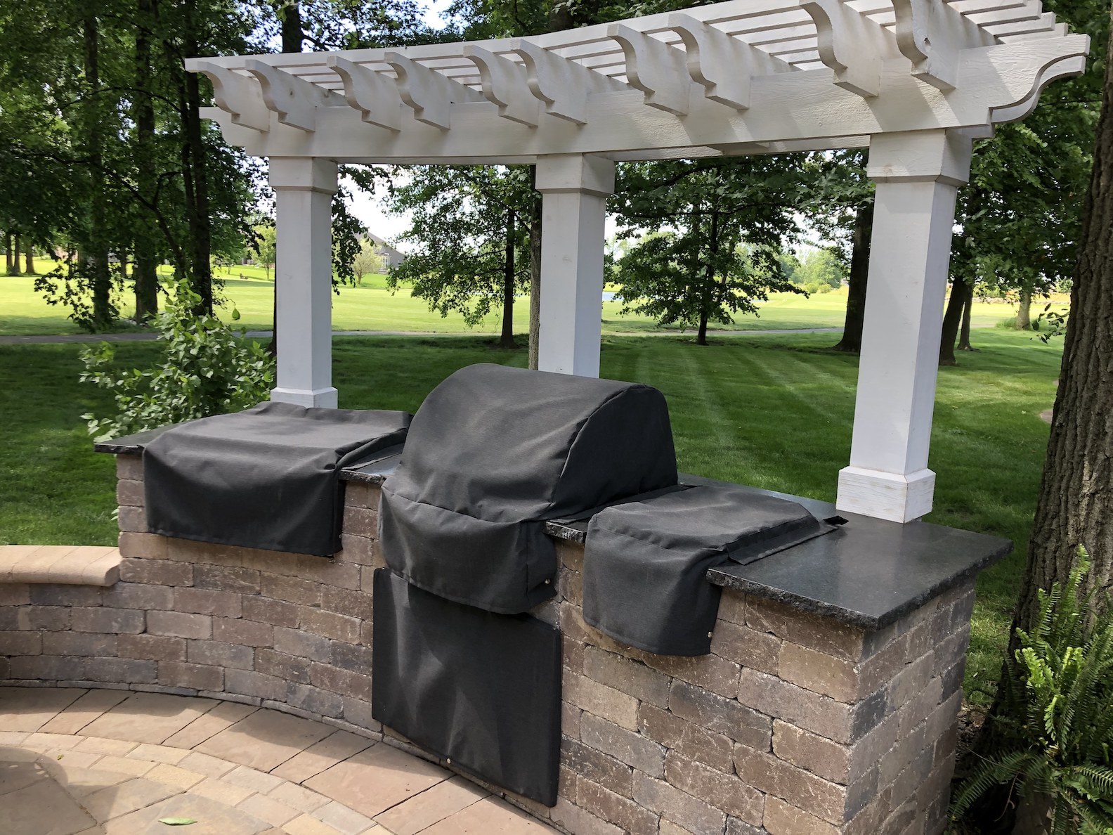 Outdoor kitchen with a stainless steel grill, burners, and sink set on a stone counter with custom fabric cover